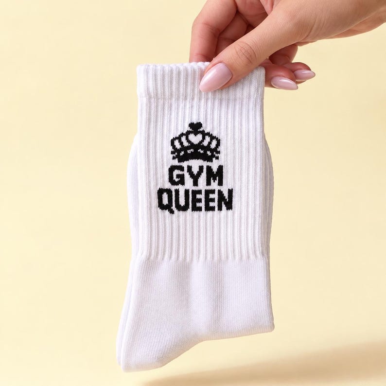 May include: A white ribbed crew sock featuring a black embroidered graphic of a crown positioned above the text "GYM QUEEN" in a bold, sans-serif font. The sock is made of a knit fabric and has a standard length with a reinforced cuff.