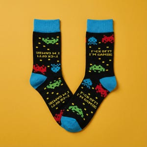 Könnte beinhalten: Ein Paar schwarze Socken mit blauen Bündchen und Zehen. Die Socken zeigen pixelige Alien-Grafiken in Rot, Grün und Blau, zusammen mit dem Text "FACK OFF! I'M GAMING" in Gelb. Die Socken sind für Gamer gedacht.