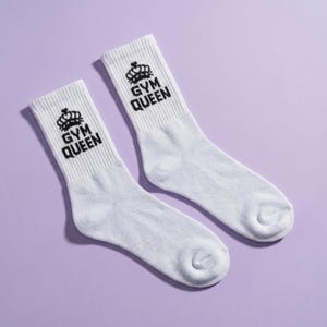 May include: A pair of white crew socks featuring a black graphic of a crown above the text "GYM QUEEN" printed on the ribbed cuff. The socks are made of a knit fabric and have a standard length that reaches above the ankle with a reinforced toe.
