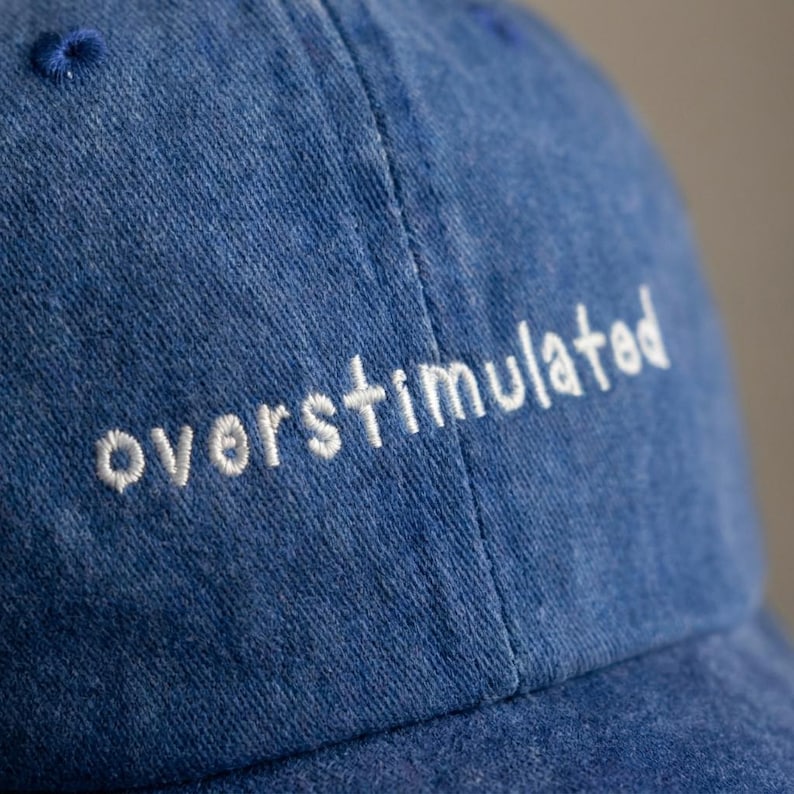 Puede incluir: Gorra de b&eacute;isbol azul denim con la palabra "overstimulated" bordada en blanco. La gorra tiene una visera curva y un peque&ntilde;o detalle cosido en la parte superior.