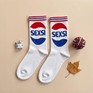 Pode incluir: Par de meias brancas com um design vermelho, branco e azul. As meias apresentam a palavra "SEXSI" em azul, com um gráfico vermelho e azul. A parte superior das meias tem listras vermelhas e azuis. Um pequeno gato de cerâmica, uma bola de lã e uma folha também estão na imagem.