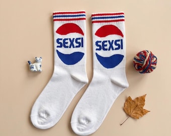 May include: Pair of white crew socks with a red, white, and blue design. The socks feature the word "SEXSI" in blue, with a red and blue graphic. The top of the socks have red and blue stripes. A small ceramic cat, a ball of yarn, and a leaf are also in the image.