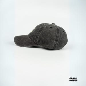May include: A charcoal gray baseball cap with a curved brim. The cap has a distressed, worn look and a small, yellow detail on the front. The cap is made of a soft, textured material and has a classic six-panel design.
