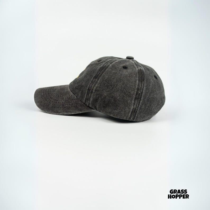 May include: A charcoal gray baseball cap with a curved brim. The cap has a distressed, worn look and a small, yellow detail on the front. The cap is made of a soft, textured material and has a classic six-panel design.