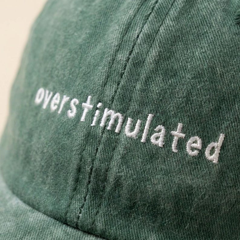 Puede incluir: Gorra de b&eacute;isbol verde con el texto bordado "overstimulated" en blanco. La gorra est&aacute; hecha de una tela texturizada y tiene una visera curva. El dise&ntilde;o es simple y moderno.