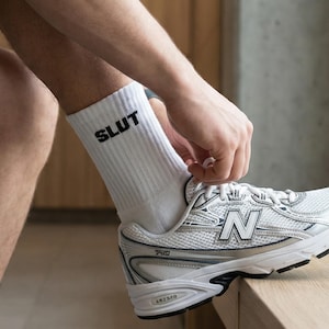 Könnte beinhalten: Eine Person bindet die Schnürsenkel eines weißen und silbernen Sportschuhs. Die Person trägt weiße Socken mit dem schwarzen Aufdruck "SLUT". Der Schuh hat den Buchstaben "N" auf der Seite.