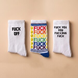 May include: Three white crew socks. The left sock features the black text FUCK OFF. The middle sock displays a rainbow-colored pattern of the word FUCK repeated vertically alongside yellow smiley faces. The right sock features the black text FUCK YOU YOU FUCKING FUCK.