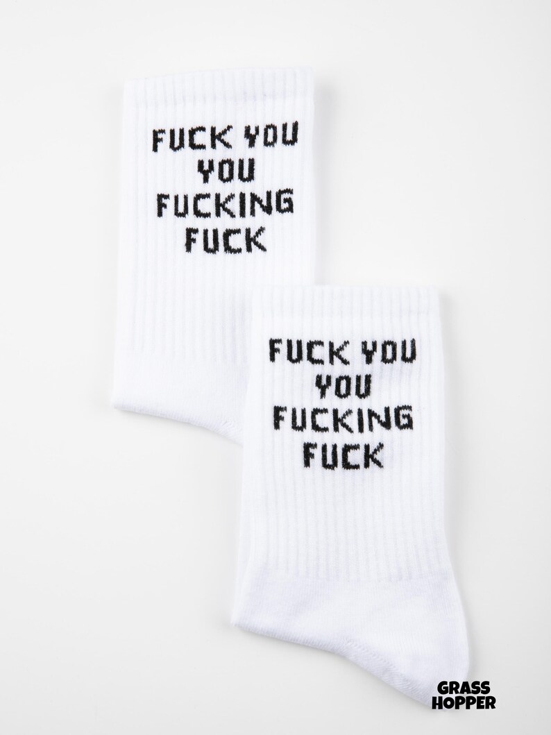 May include: Two white socks with the text "FUCK YOU YOU FUCKING FUCK" printed in black. The socks are ribbed and have a casual style. The brand name "GRASSHOPPER" is printed in the bottom right corner.
