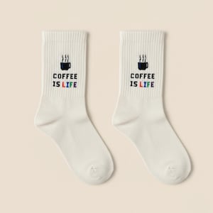 Könnte beinhalten: Ein Paar weiße Socken mit dem Aufdruck "COFFEE IS LIFE" in Schwarz, Blau, Rot und Grün. Oberhalb des Textes befindet sich eine schwarze Kaffeetasse mit Dampf. Die Socken sind gerippt.