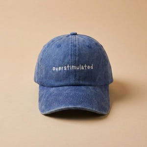 May include: A blue denim baseball cap with the word "overstimulated" embroidered in white. The cap has a curved peak and a classic six-panel design. The hat is a solid blue colour and has a slightly faded appearance.