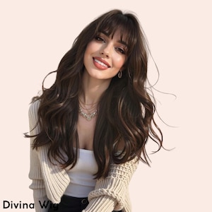 Puede incluir: Una mujer con cabello largo, ondulado y castaño oscuro con flequillo. Lleva una rebeca color crema sobre una camiseta blanca y un delicado collar. El texto "Divina Wig" es visible en la parte inferior.