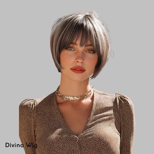 Puede incluir: Una mujer con una peluca corta y en capas con tonos grises y marrones y un flequillo recto. Lleva un collar dorado y una blusa marrón estampada. El texto "Divina Wig" es visible en la parte inferior izquierda.
