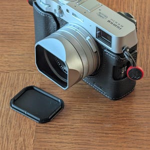 Könnte beinhalten: Silberne Fujifilm X100V Digitalkamera mit schwarzem Lederetui und Sonnenblende. Die Kamera hat mehrere Drehknöpfe und Tasten, ein Objektiv und einen schwarzen Objektivdeckel. Auf einer Holzoberfläche.