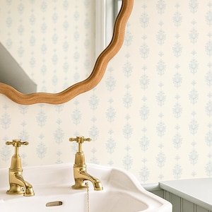 Op de afbeelding: Badkamerinterieur met een witte wastafel en gouden kranen. Boven de wastafel hangt een houten, geschulpte spiegel, geflankeerd door twee wandkandelaars met bruine tinten. De muren zijn voorzien van behang met patronen en geschilderde lambrisering.