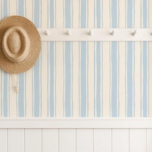 May include: A wall covered in wallpaper featuring vertical light blue and cream stripes. A white wooden peg rail is mounted on the wall, holding a tan straw hat. Below, a pair of tall black rubber boots stands against white vertical wainscoting.