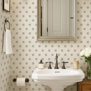 Puede incluir: Un baño pequeño con un lavabo con pedestal, espejo y mueble de madera. Las paredes están cubiertas con papel pintado con un patrón repetitivo de círculos azul-gris. Dos apliques flanquean el espejo y un jarrón con flores blancas se encuentra sobre el mueble.