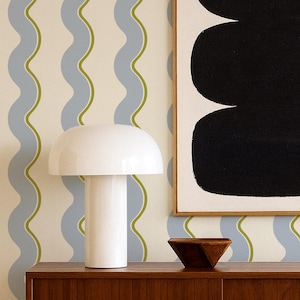 Puede incluir: Un salón de estilo moderno de mediados de siglo. Cuenta con un mueble de madera oscura, una lámpara de seta blanca y una pintura abstracta con tres óvalos negros. El papel pintado tiene un patrón ondulado azul y verde.