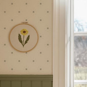 Puede incluir: Un aro de bordado redondo con una flor amarilla y hojas verdes. La obra de arte está colgada en una pared con papel pintado con estampado de estrellas y un panel verde. Se ve una ventana con una cortina blanca.