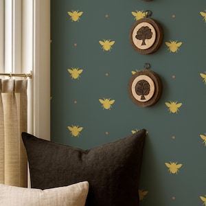 May include: An interior scene with a dark teal wall decorated with gold bee and dot patterns. Three wooden circular wall hangings with tree designs are displayed. Two pillows, one beige and one dark brown, rest on a wooden surface.