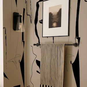 May include: A black and white striped towel hangs on a matte black metal towel bar mounted to a beige wall featuring a black abstract line pattern. Above the towel is a framed black and white landscape photograph and a black wall sconce light.