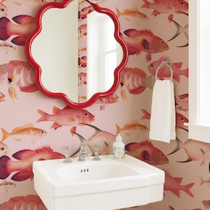 May include: A white pedestal sink with a chrome faucet and a soap dispenser, positioned against wallpaper featuring a repeating pattern of pink and orange fish. A red scalloped mirror hangs above the sink, and a white towel hangs on a ring.