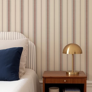 Può includere: Scena di camera da letto con un comodino in legno, un cassetto e una mensola aperta. Sopra c'è una lampada dorata a forma di cupola. Un cuscino blu navy poggia su un letto bianco, con carta da parati a righe sullo sfondo.