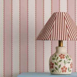 May include: A decorative table lamp with a red and white striped lampshade sits on a light blue cabinet. The lamp base is cream-colored with red floral designs. The cabinet holds books and a small jar.