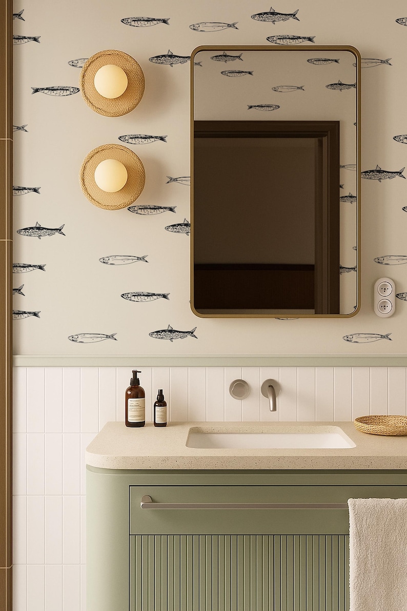 K&ouml;nnte beinhalten: Badezimmer mit einer salbeigr&uuml;nen Waschtischunterschrank, einem wei&szlig;en Waschbecken und einem goldgerahmten Spiegel. Die Wand ist mit einer Tapete mit Fischmuster verziert. Zwei geflochtene Wandleuchten und ein Seifenspender sind ebenfalls zu sehen.