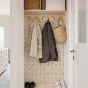 May include: A closet storage area featuring a light wood shelf with five wooden hooks, a woven basket, a beige trench coat, a dark gray coat, a straw tote bag, and a pair of black shoes and white sneakers on a lower light wood shelf.