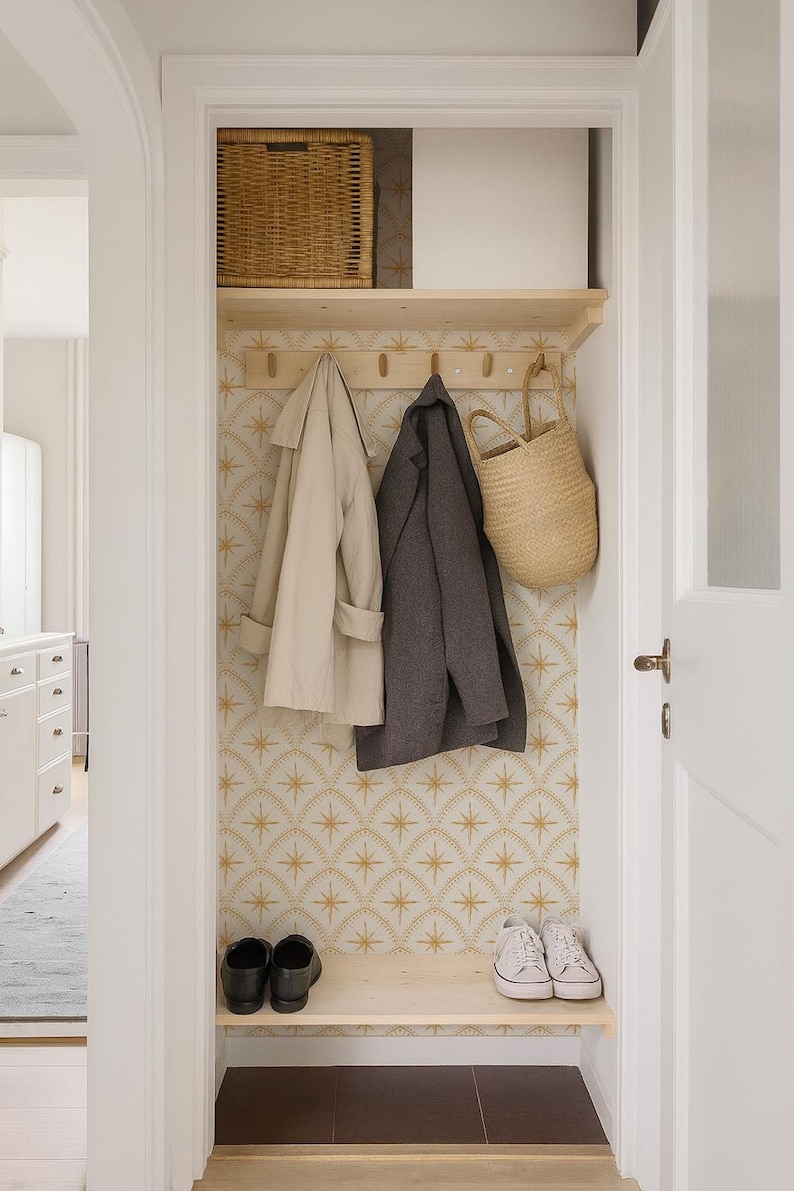 May include: A closet storage area featuring a light wood shelf with five wooden hooks, a woven basket, a beige trench coat, a dark gray coat, a straw tote bag, and a pair of black shoes and white sneakers on a lower light wood shelf.