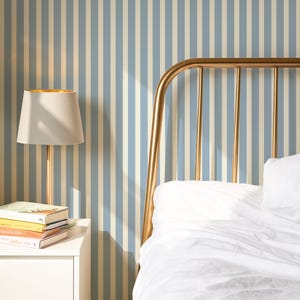 May include: A bedroom interior with a gold-coloured bed frame and white bedding. A white bedside table holds a lamp with a gold interior, books, and a small potted plant. The wall has a blue and white striped pattern.