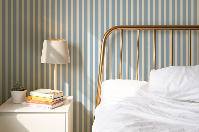 May include: A bedroom interior with a gold-coloured bed frame and white bedding. A white bedside table holds a lamp with a gold interior, books, and a small potted plant. The wall has a blue and white striped pattern.