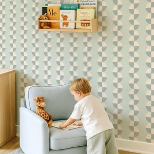 May include: A child's room with a light blue armchair, a giraffe stuffed animal, and a child looking at a book. The room has a light green and white patterned wallpaper and wooden bookshelves with children's books.