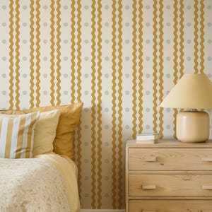 May include: A bedroom scene featuring a bed with yellow bedding and striped pillows. The wall is decorated with a cream-colored wallpaper with vertical wavy mustard-yellow stripes and small gray floral accents. A wooden nightstand and lamp complete the scene.