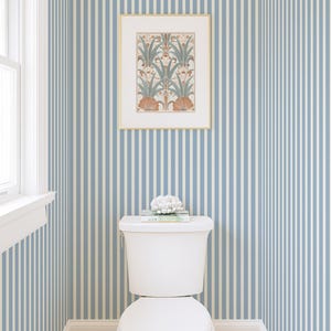 May include: A white toilet in a bathroom with blue and white striped wallpaper. A framed botanical print hangs above the toilet. A decorative object and books sit on the toilet cistern. The floor has a pink and white patterned tile.