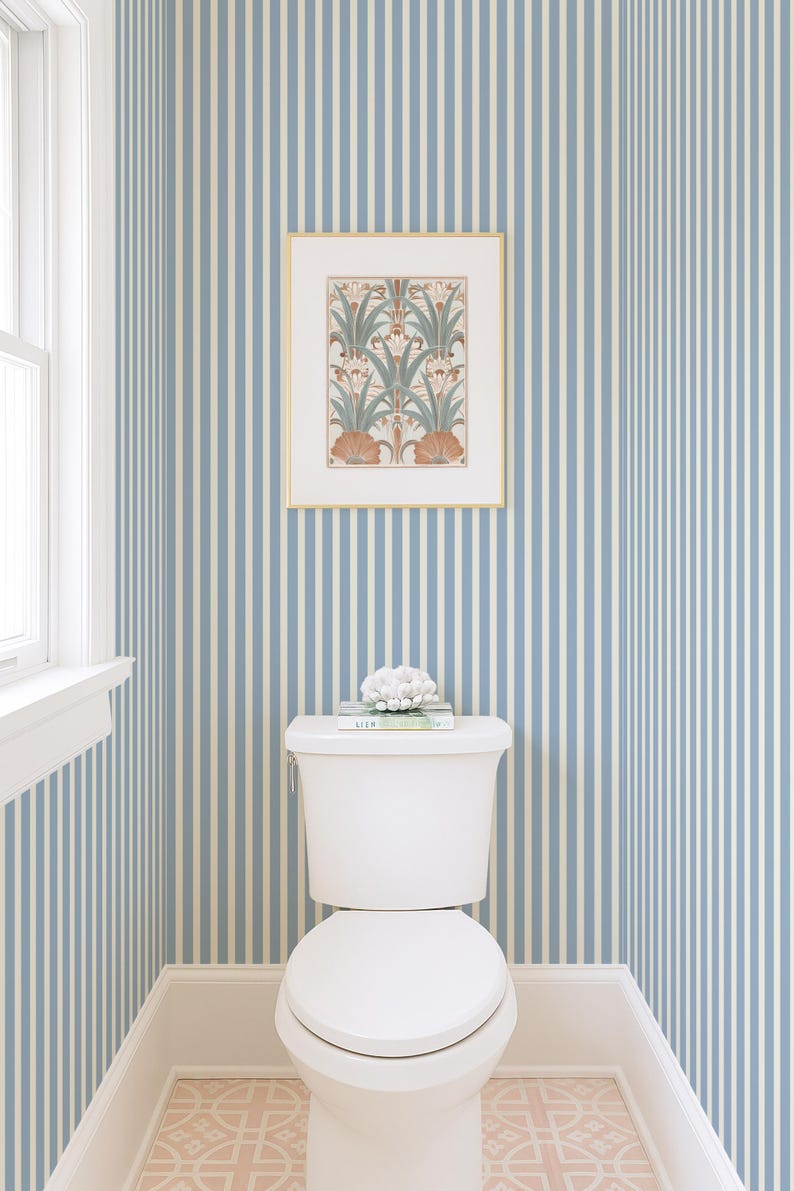 May include: A white toilet in a bathroom with blue and white striped wallpaper. A framed botanical print hangs above the toilet. A decorative object and books sit on the toilet cistern. The floor has a pink and white patterned tile.
