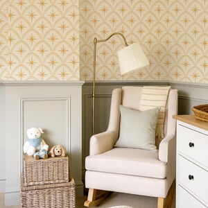 May include: A beige upholstered rocking chair with a light green throw pillow and a striped blanket, positioned near a brass floor lamp, two stacked wicker storage baskets with plush toys, a white dresser, and a cream-colored knit pouf.