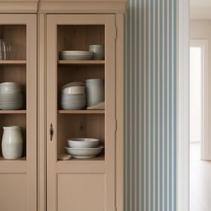 May include: A beige cabinet with glass doors displaying white crockery and ceramic items. A woven basket sits on top. The background features blue and white striped wallpaper, with a doorway on the right.