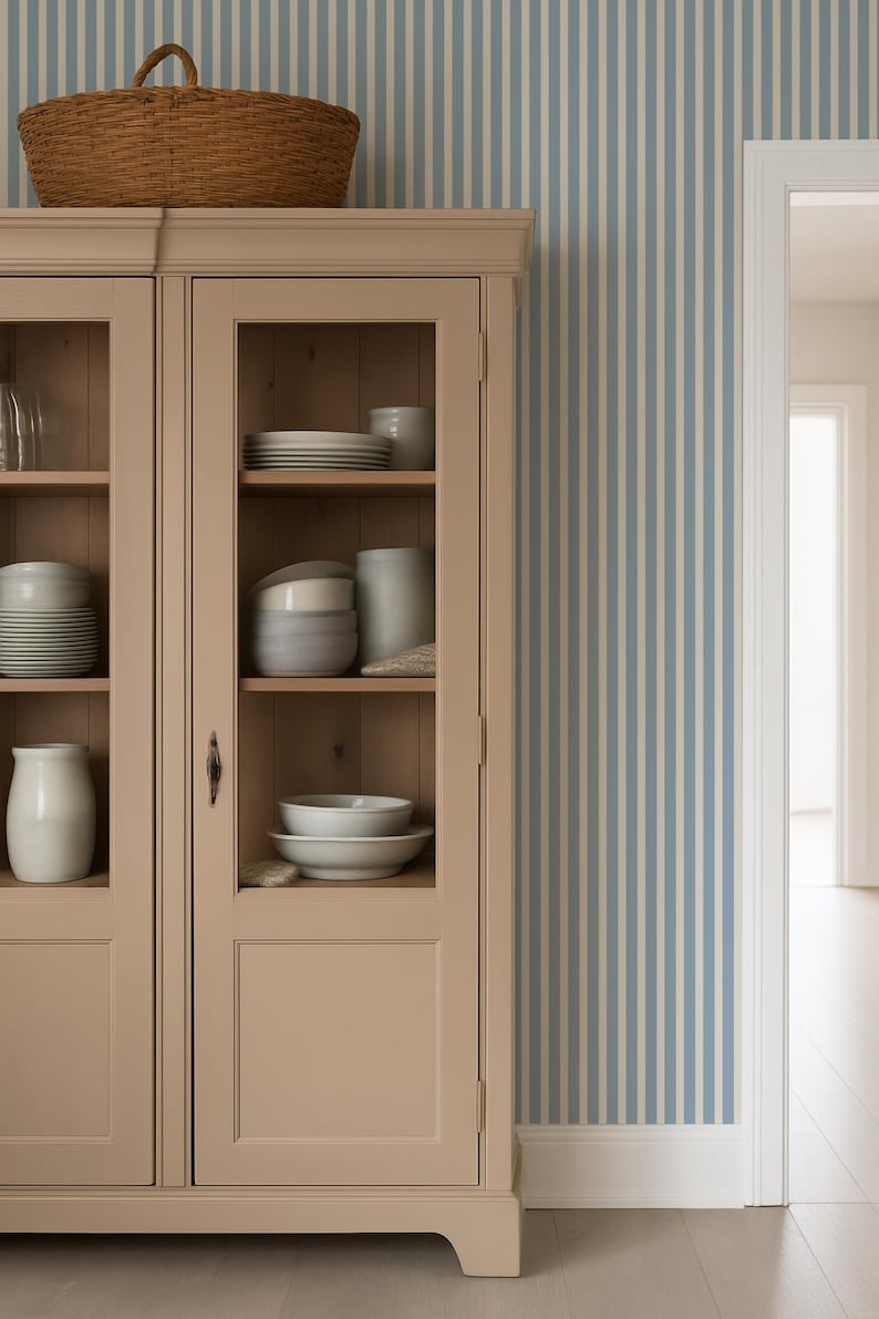 May include: A beige cabinet with glass doors displaying white crockery and ceramic items. A woven basket sits on top. The background features blue and white striped wallpaper, with a doorway on the right.