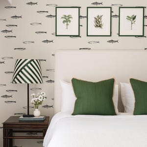 K&ouml;nnte beinhalten: Schlafzimmer-Interieur mit einem Bett mit wei&szlig;er Bettw&auml;sche und gr&uuml;nen Kissen. Die Wand ist mit einem Fischmuster und gerahmten botanischen Drucken verziert. Eine gestreifte Lampe und ein Nachttisch aus dunklem Holz sind ebenfalls zu sehen.