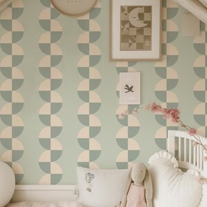 May include: A nursery scene with a light green and cream geometric patterned wallpaper. A framed picture of a moon and stars hangs on the wall. A stuffed bunny and heart-shaped pillow rest on a bed with a pink blanket.