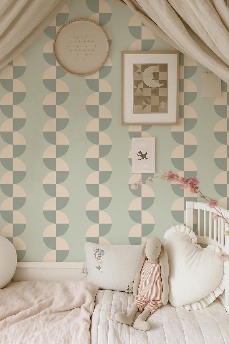 May include: A nursery scene with a light green and cream geometric patterned wallpaper. A framed picture of a moon and stars hangs on the wall. A stuffed bunny and heart-shaped pillow rest on a bed with a pink blanket.
