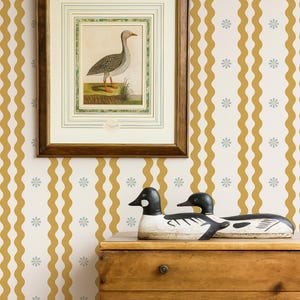 May include: A framed bird illustration hangs above a wooden dresser, with two decorative duck decoys. The background features a wallpaper pattern of wavy gold stripes and small blue flowers. The dresser has two drawers.