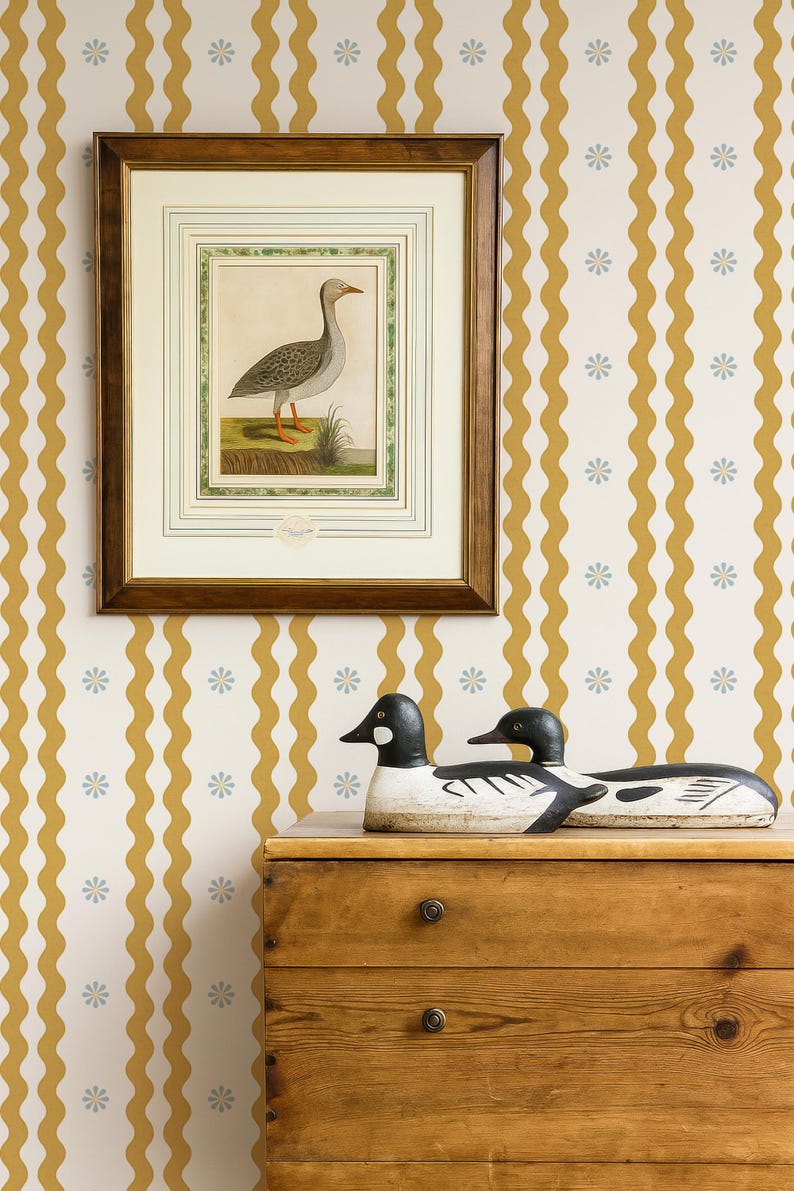 May include: A framed bird illustration hangs above a wooden dresser, with two decorative duck decoys. The background features a wallpaper pattern of wavy gold stripes and small blue flowers. The dresser has two drawers.
