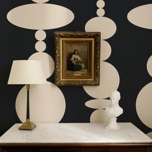Pode incluir: Uma c&oacute;moda de madeira escura com tampo de m&aacute;rmore branco, um candeeiro de mesa com abajur creme, uma pintura a &oacute;leo emoldurada de uma figura sentada e um busto branco. A parede tem papel de parede preto com um padr&atilde;o de grandes c&iacute;rculos creme.