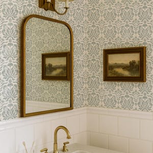 May include: A bathroom vanity area featuring a white marble countertop with a brass tap, a glass cup with two toothbrushes, a brass-framed arched mirror, a double-light wall sconce, and two landscape paintings against blue floral wallpaper.
