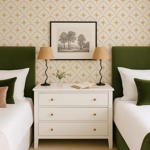 May include: A white three-drawer dresser with gold knobs sits between two beds with green velvet headboards. Two lamps with wavy black bases and pleated tan shades flank a framed black and white landscape print on the patterned wallpaper.