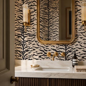 Pode incluir: Um móvel de casa de banho com bancada de mármore branco, armário canelado castanho escuro e torneira dourada. A parede está coberta com papel de parede bege com um padrão de riscas de tigre pretas. Um espelho dourado pende sobre o lavatório.