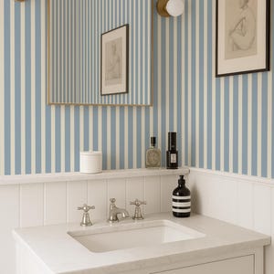 May include: A bathroom interior with a white marble vanity and chrome taps. The walls are decorated with blue and white striped wallpaper. A framed picture and a mirror are mounted above the vanity, alongside a shelf with toiletries.