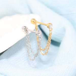 Puede incluir: Dos piercings industriales tipo barbell, uno en tono plateado y otro en tono dorado. Cada barbell presenta una barra metálica recta con una piedra preciosa redonda y transparente en un extremo y una doble cadena colgante que une ambos lados.